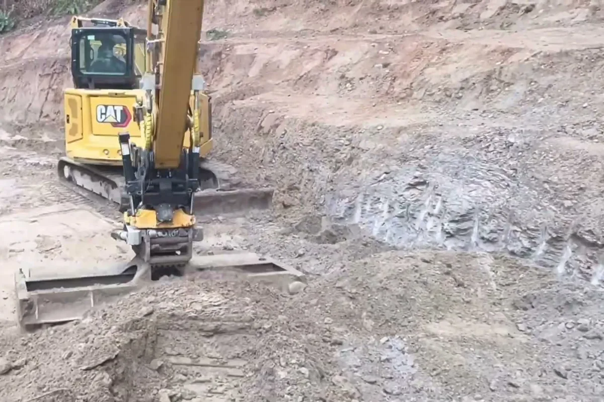 Mini excavator with a tilt rotator cutting a level bench into a slope before a retaining wall goes in